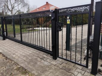 Czarna kuta brama przesuwna z furtką ogrodzeniową na tle domu z czerwonym dachem