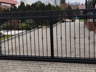 Czarna kuta brama przesuwna z ozdobnym wzorem, wjazd na kostkę brukową przed domem