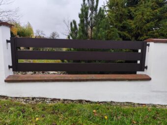 Nowoczesne ogrodzenie aluminiowe czarne, poziome panele na białym murowanym słupku, w tle drzewa